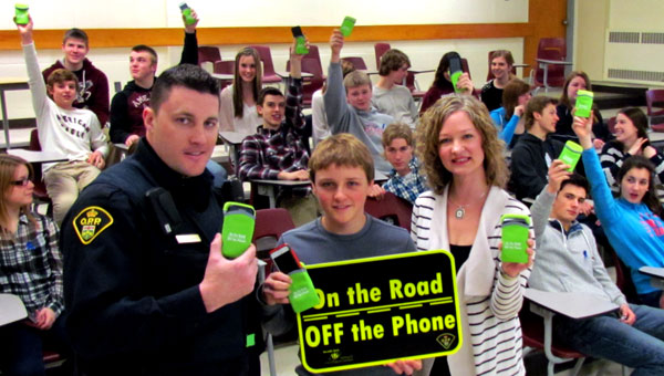 Constable Anthony Mann with PECI student Tyler Allison and the health unit's Kerri Jianopoulos show off the new On the Road, Off the Phone signs to be placed on roads around the school this spring. In the background, students participating in the presentation hold up phone covers designed as reminders to put their phones away when driving.